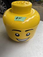 Large yellow LEGO storage head with smiling face and 'LEGO' embossed lid, placed on table outdoors.