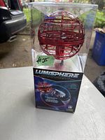 Front view of the unopened Lumisphere orb in its clear plastic packaging showing the red spherical orb with grid design and black base.