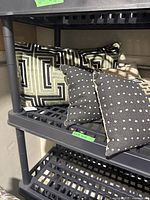 Three cushions on a black shelf: one large cushion with black and beige geometric maze-like pattern, two smaller black cushions with white polka dots.