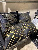 Four black velvet cushions and pillows with gold abstract and geometric patterns, stacked on a shelf.