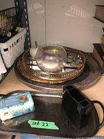 Stack of decorative metal trays with embossed gold and bronze tone patterns, clear glass candle holder, bar of soap, and seashells on a shelf.