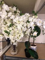 Three potted faux flower arrangements on a table: large white floral bouquet in glass vase filled with white pebbles, white orchid in black pot, green topiary in decorative pot.