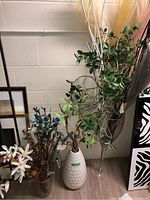 White textured ceramic vase with faux leafy branches and white sticks, clear tall glass vase with faux greenery and pampas grass, and a smaller clear glass vase with dried brown botanical pods.