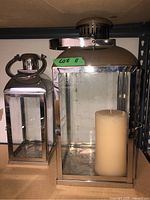 Two metal framed lanterns shown side by side on a shelf, one containing an ivory pillar candle. Both have glass panels and chrome-like finish.