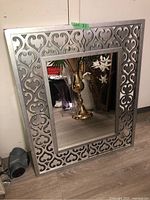 Front angled shot of decorative mirror showing the ornate scrollwork patterned MDF frame painted in metallic silver and the clear central mirror pane.