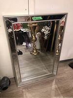 Front view of a rectangular beveled mirror with a silver-toned beaded frame, resting against a wall and reflecting interior furnishings including a brass vase.