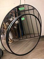 Two wall mirrors with black metal frames leaning against a wall on a wood floor. One is round and the other arched. The round mirror is smaller in diameter than the arched one.