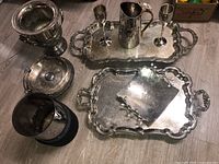 View of silver plate servingware including trays, goblet glasses, vase, and lidded container laid out on floor showing scale and condition.
