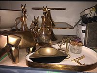 Photo of assorted gold-tone servingware and decor items including geometric shapes, birds, and a small decorative jar on a flat white tray.