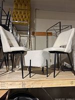 Two white upholstered chairs with black legs and a matching round upholstered stool with black legs displayed on a wooden platform indoors.