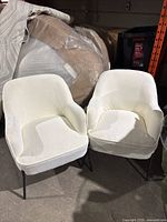 Two white fabric chairs with metal legs placed side by side on concrete floor, with some packaging and materials in the background.