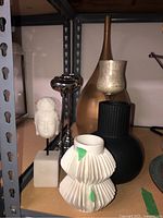 Multiple decorative vases and candleholders arranged on a shelf, showing variety of materials and finishes including metallic gold, black ribbed, white pleated ceramic, white bust sculpture, and silver candleholders.