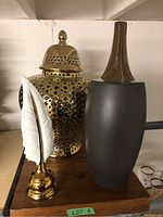 Three decorative items shown together on wooden tray: gold-tone metal lidded vase with perforated pattern, tall ceramic vase with bronze neck, white ceramic feather pen holder on gold base.
