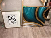 Two framed wall art pieces against a wall, one smaller white-framed abstract dot pattern print and one larger gold framed abstract teal and gold painting.