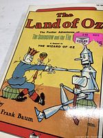 Front cover of The Land of Oz paperback book showing characters Scarecrow and Tin Woodman shaking hands, authored by L. Frank Baum.