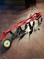 Full view of the red metal toy bottom plow with four black plowshares and single front wheel.