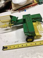 Side angle view of green and yellow Ertl John Deere hay baler toy on white surface with measuring tape showing approximately 7 inches length.
