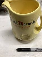 Front angle view of yellow ceramic pitcher with Grand Macnish Scotch Whisky logo, with Sharpie pen for scale.