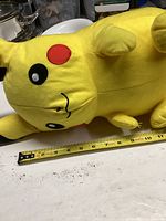 Side view of plush Pikachu lying on table with a measuring tape showing length about 17 inches, showing face and part of the body.