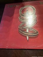 Front cover of the Toto IV album featuring a sword piercing four rings on a red background with the band name 'TOTO' in gold letters.