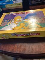 Box cover showing Homer and Marge Simpson arm wrestling, with the game title 'Battle of the Sexes'