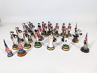 Wide view of multiple Revolutionary War themed chess pieces arranged in rows, depicting soldiers in red and blue coats, flag bearers, and mounted cavalry figures on circular bases.