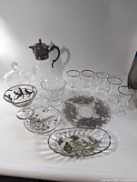 Overview of the lot showing the Godinger carafe with silver lid and handle, six glasses with silver rims, and various plates and bowls with silver floral designs on clear glass.
