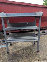 Front view of gray wooden plant stand showing three slatted shelves and metal hooks on sides, placed outdoors in front of red container.