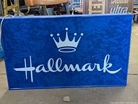Full front view of rectangular blue metal lighted Hallmark sign with white logo and script, power cord draped on top.