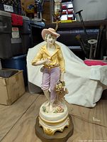 Front view of tall Victorian style ceramic lamp base depicting a boy holding a basket of fruit and flowers, with pink pants and yellow jacket, standing on an ornate base with gold accents. Lamp hardware seen at top with bulb socket, no shade attached.