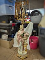 Full view of tall Victorian style figural lady and cherub candelabra lamp. The porcelain/resin base depicts a draped lady holding flowers next to cherub and topped with a multi-arm gold candelabra. Damage to shade noted but shade not included.