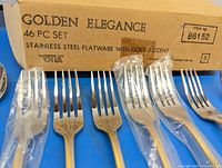 Photo showing the original cardboard box labeled 'Golden Elegance 46 PC Set Stainless Steel Flatware with Gold Accent' by Stia and some forks wrapped in plastic.