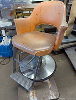 Side angle view of vintage barber chair showing orange padded vinyl upholstery, metal base and footrest. Shows wear and dirt.