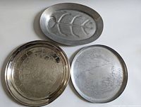 Three metal trays shown, one oval with fishbone design, two round engraved with detailed landscapes
