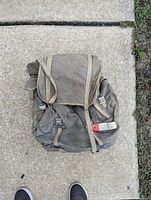 Front view of heavily used vintage canvas hiking backpack lying on concrete showing the flap, buckled front pockets, and shoulder straps.
