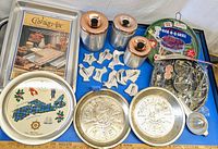 Overview photo showing all items in the lot arranged on a white surface. Contains metal canisters, pie plates, cookie cutters, souvenir trays, BBQ grill cover, and shrimp baskets.