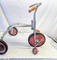 Side view showing the tricycle's silver cast aluminum frame, rubber tires with red painted rims, plastic pedals, and brown plastic handle grips.
