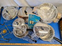 Overview of cookware set on blue table with plastic wrapping on lids and pots, showing multiple stainless steel pots and lids with black handles and knobs.