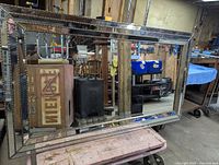 Large rectangular wall mirror with decorative silver-tone frame showing small square mirrored tiles around the edges. Mirror is positioned leaning against a wooden surface in a workshop or storage area.