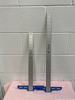 Two aluminum T-square rulers against a white wall, one 18 inch and one 24 inch, with blue plastic handle bases.