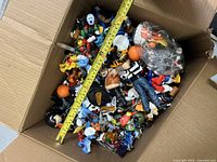 Open box filled with a variety of small character figurines and keychains from Disney, Marvel, and other brands, showing scale with measuring tape.