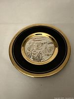 Full view of round plate with black and gold rim and embossed silver-toned center with swans design.