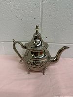 Front side view of silver plated Moroccan teapot showing handle, spout, lid, and legged base with decorative engravings.
