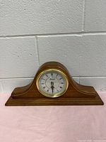 Full view of wooden mantel clock with DUMAI branding, displayed on a pink surface against a white textured wall.