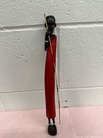 Front and side view of the Masai wood figurine, showing the red garment and spear against a white wall background.