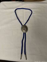 Full view of the vintage silver metal bolo tie with blue braided cord displayed on a white background.