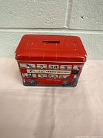 Front and side view of the vintage Churchill's tin box showing London double-decker bus graphics and coin slot on lid.