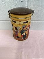 Front view of the cylindrical tin can with a brown lid, showing illustrated people at a Tim Hortons cafe sitting and conversing.