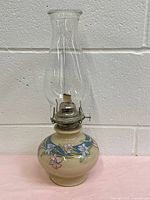 Front view of ivory oil lamp with floral painted decoration, showing clear glass chimney and metal burner against white wall background.