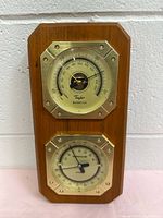 Front view of wooden framed wall mounted weather station showing upper barometer and thermometer dials with metallic octagonal bezels.